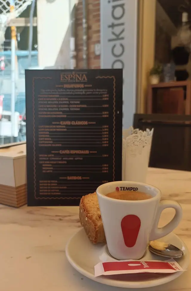 Café espresso en taza blanca y roja con un trozo de bizcocho, con la carta del local al fondo
