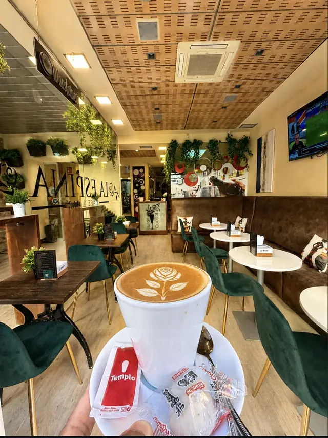 Café con una rosa dibujada en la espuma y, al fondo, el interior del local con sillas verdes y plantas